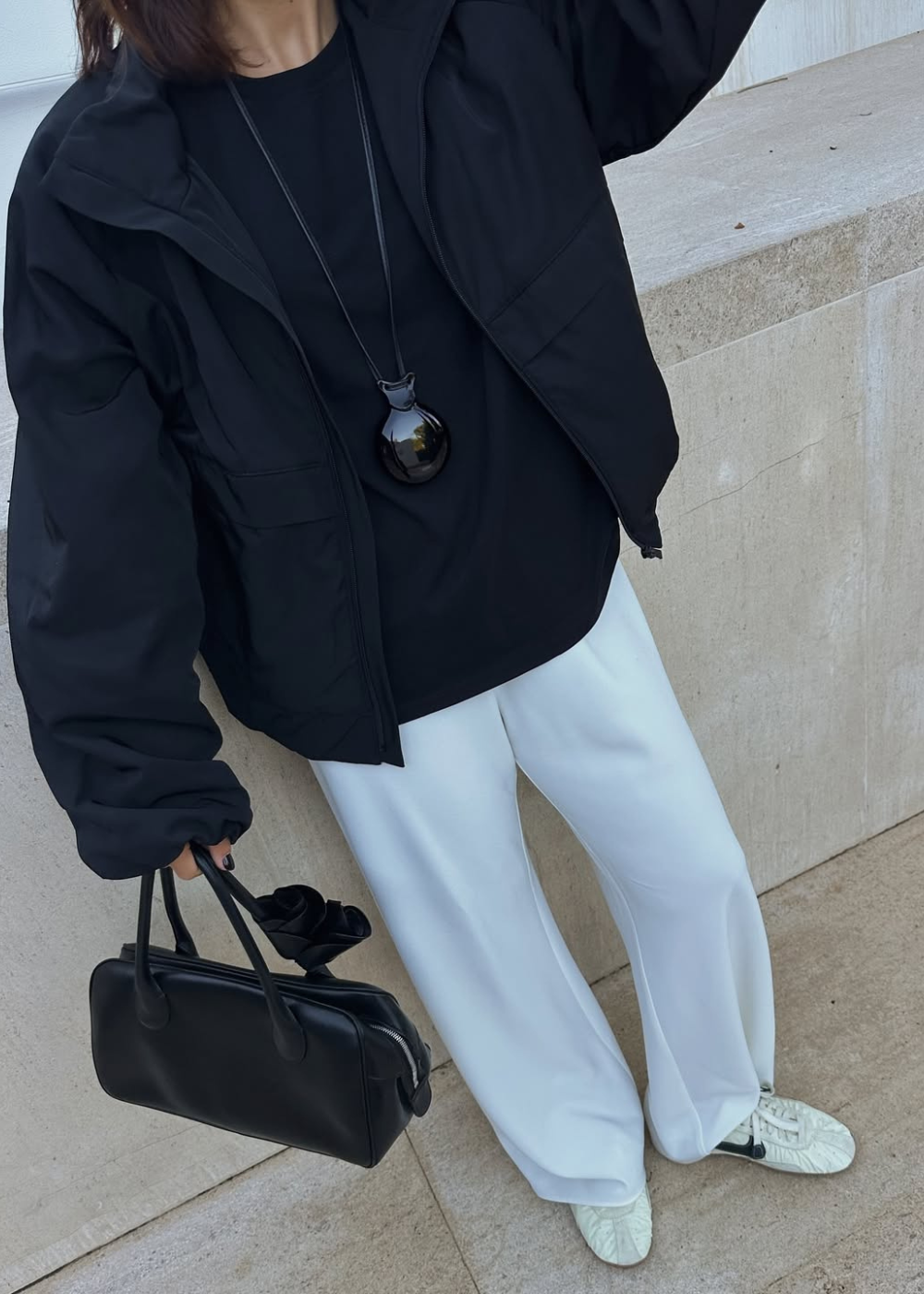 Modelo veste jaqueta preta oversized, camiseta preta, calça branca ampla e sapatos flat brancos com detalhes pretos.