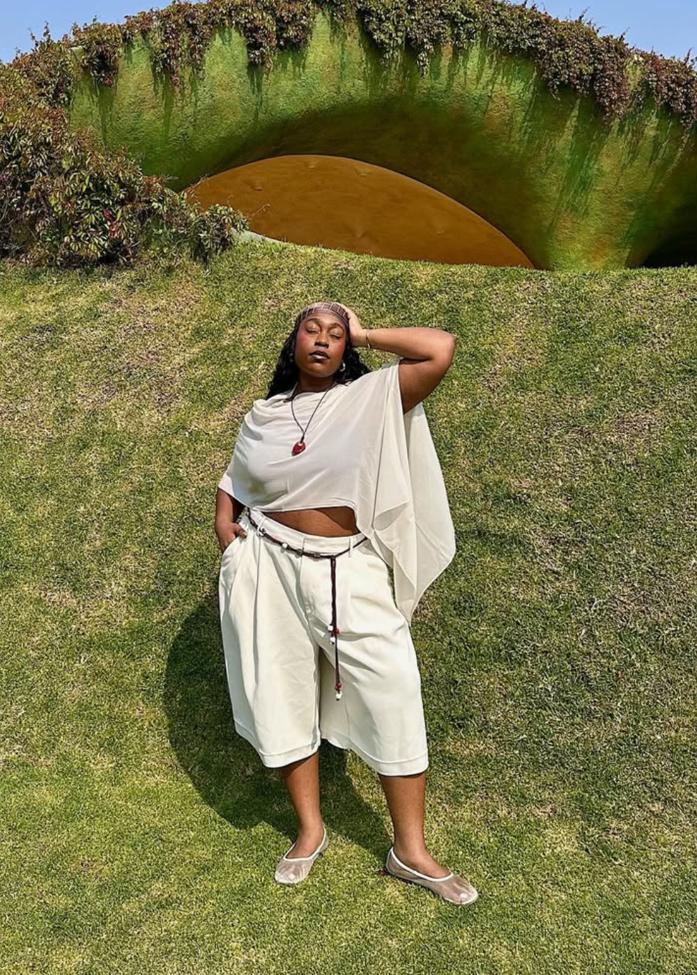 Modelo posa com postura confiante usando blusa assimétrica fluida, bermuda ampla off-white e sapatos flat transparentes.