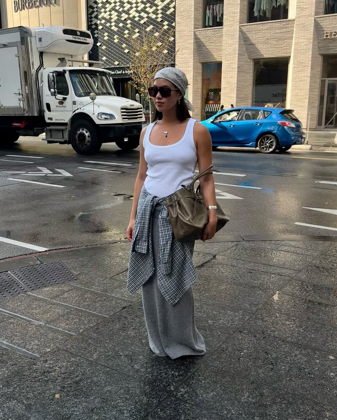 Modelo posa na rua com regatas branca básica de algodão, calça cinza larga, camisa xadrez amarrada na cintura e lenço na