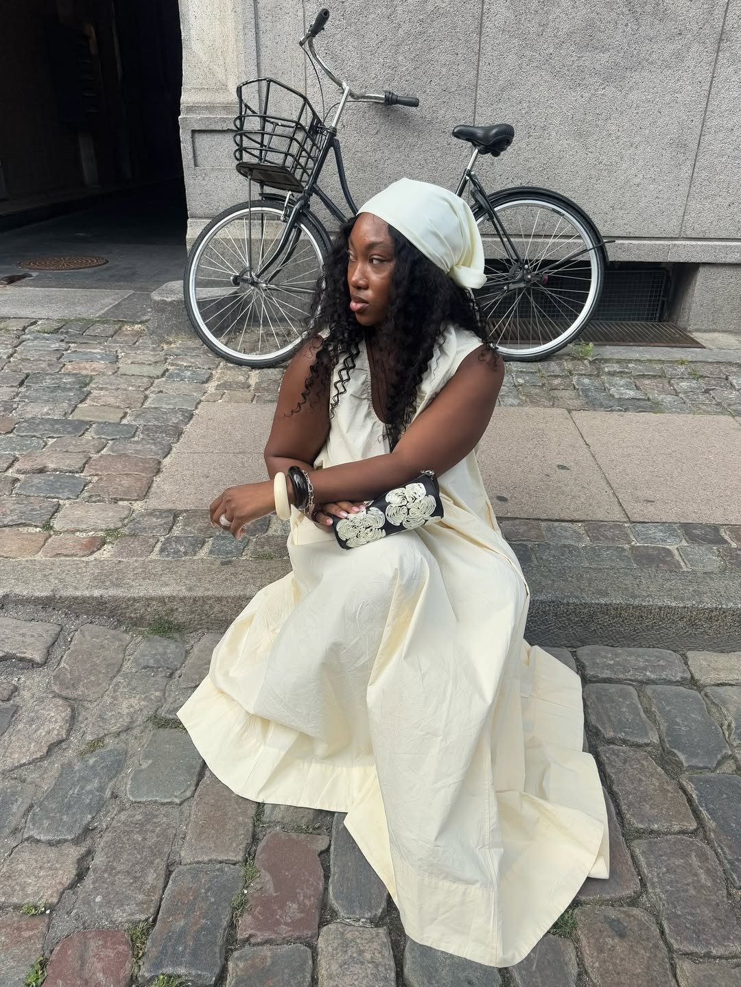 Modelo senta no chão de pedra com vestido regata longo e fluido em tom creme, combinando lenço e acessórios para look suave