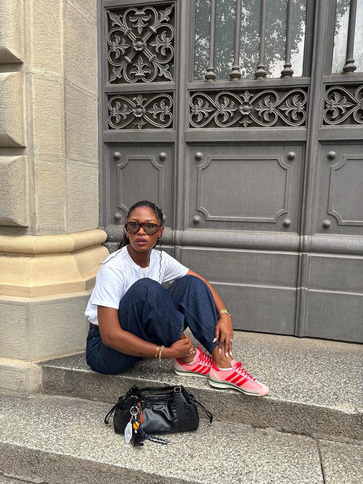 Modelo senta casualmente em degrau urbano, usando sneaker feminino rosa com listras vermelhas, calça jeans escura e camiseta