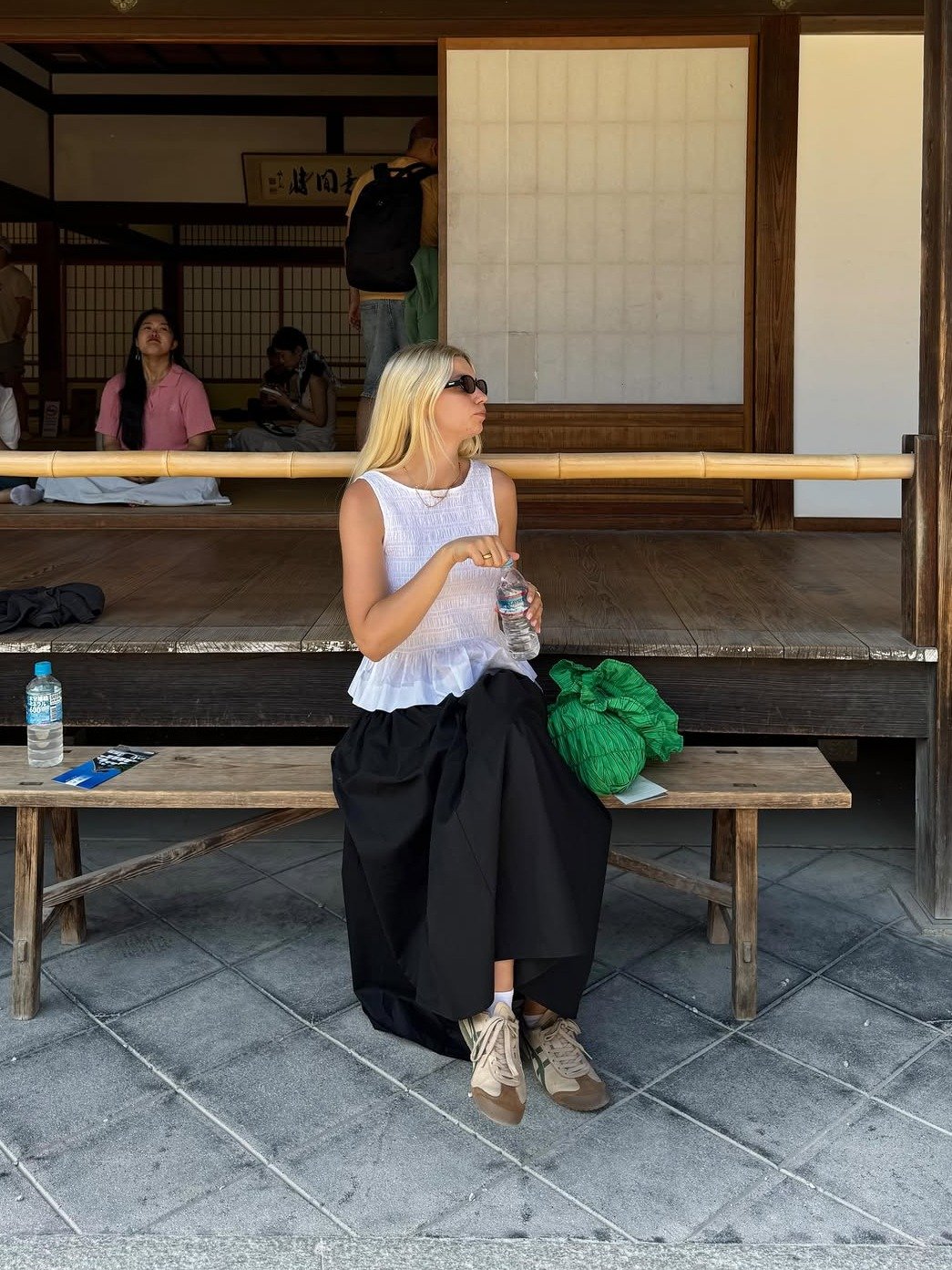 Mulher sentada usa sneaker feminino bege com detalhes em verde, saia preta ampla e blusa branca sem mangas, em clima