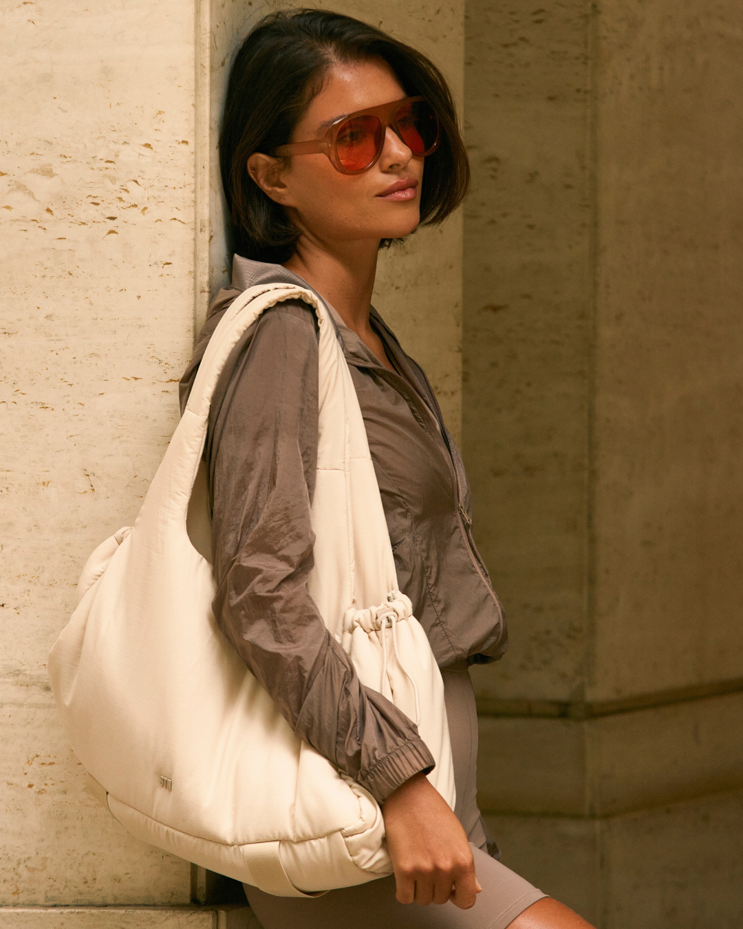Modelo posa encostada com jaqueta nylon marrom, bermuda bege justa e bolsa oversized off-white, tendência athleisure