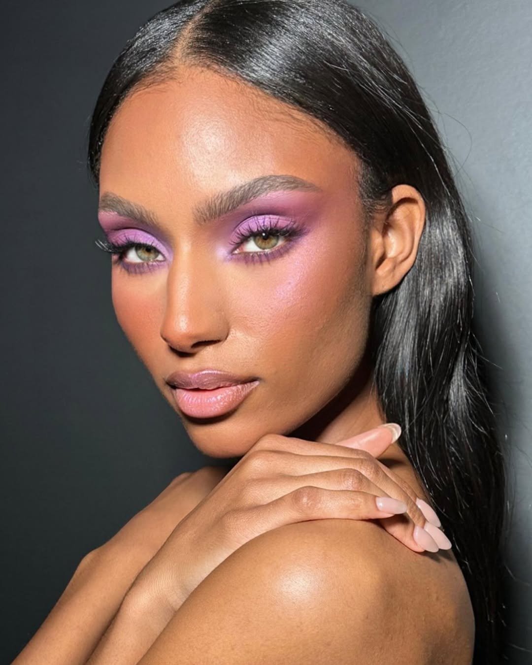 Modelo posa com maquiagem vibrante em tons de lilás e rosa, pele iluminada e expressão serena, destacando tendências