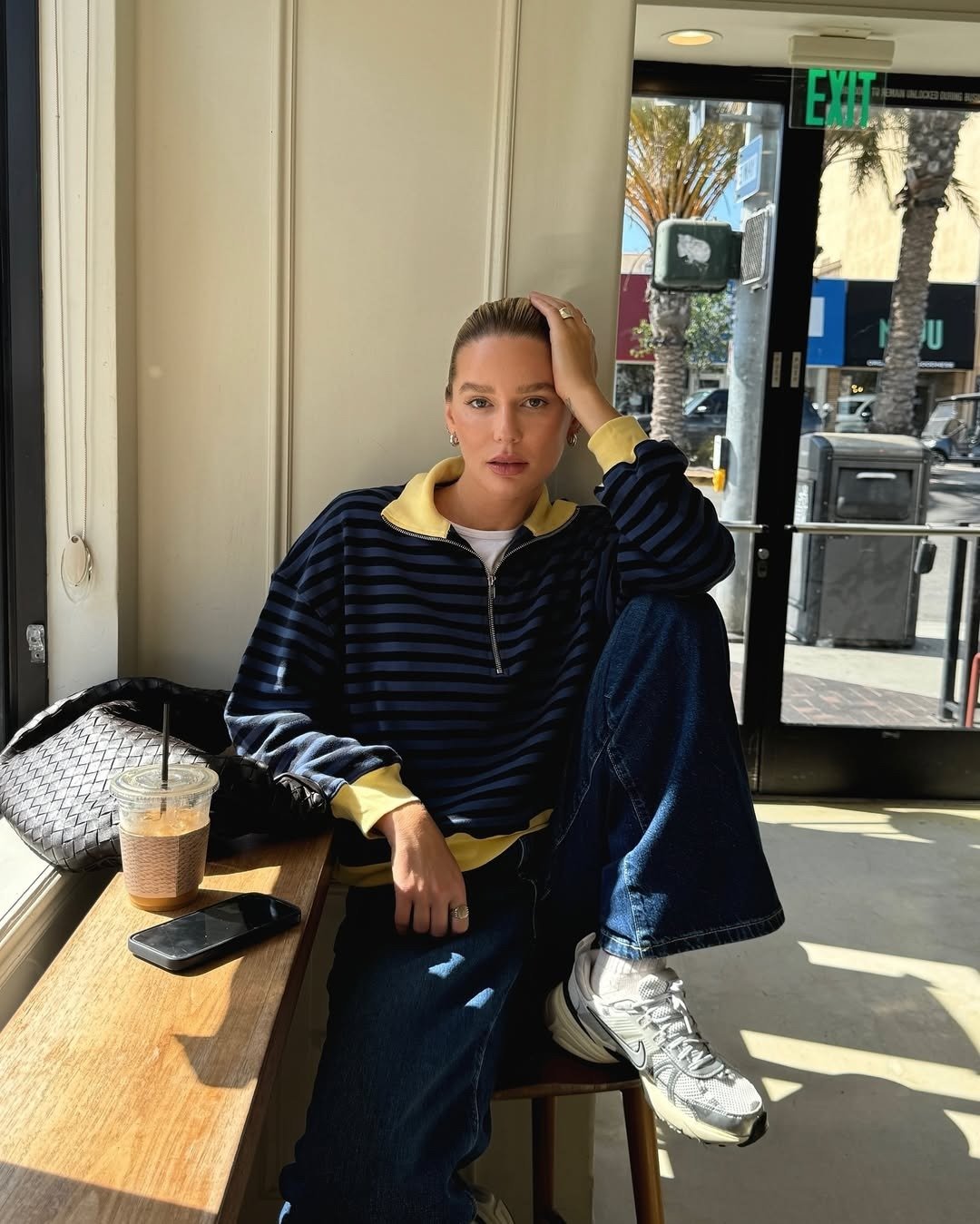 Modelo posa sentada com tênis retrô branco de design robusto, calça jeans ampla e moletom listrado azul-marinho com detalhes