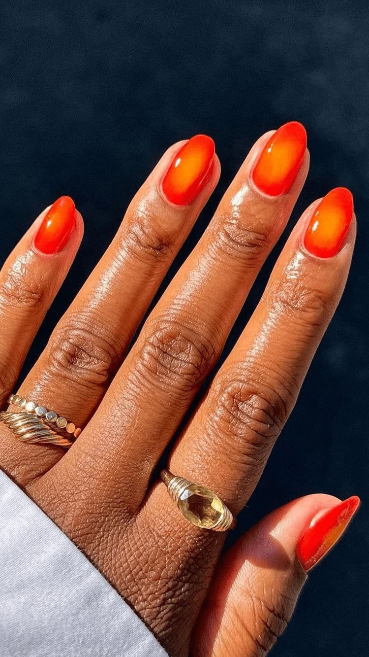 Unhas coloridas da moda em degradê laranja vibrante com acabamento brilhante destacam dedos adornados por anéis dourados