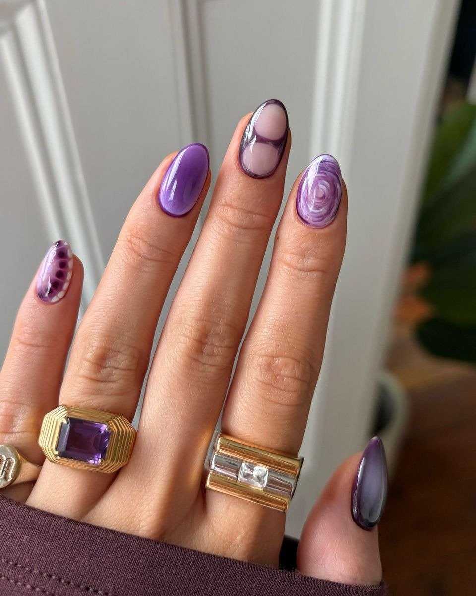 Unhas coloridas da moda em tons de roxo com desenhos abstratos e acabamento glossy, destacando anéis dourados e prata para