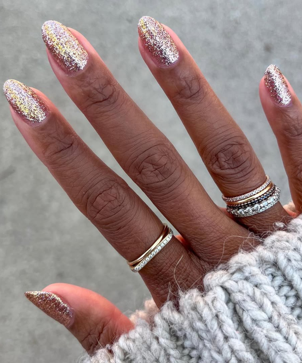 Unhas para o Carnaval com glitter dourado e prata em formato oval, combinando anéis delicados e suéter de tricô cinza claro.
