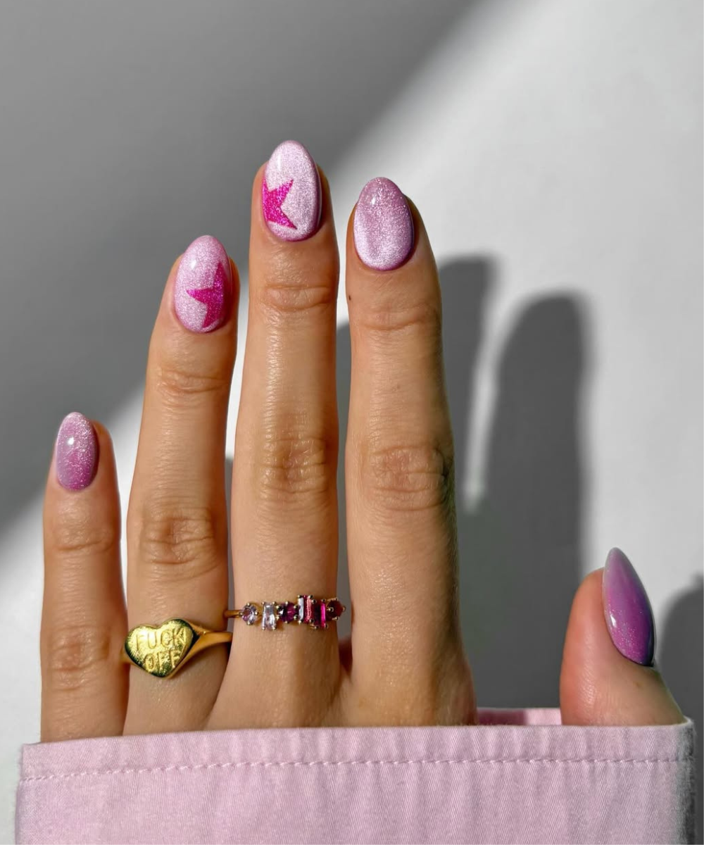 Unhas para o Carnaval em tom rosa cintilante com estrelas fúcsia, formato oval e acessórios dourados delicados.