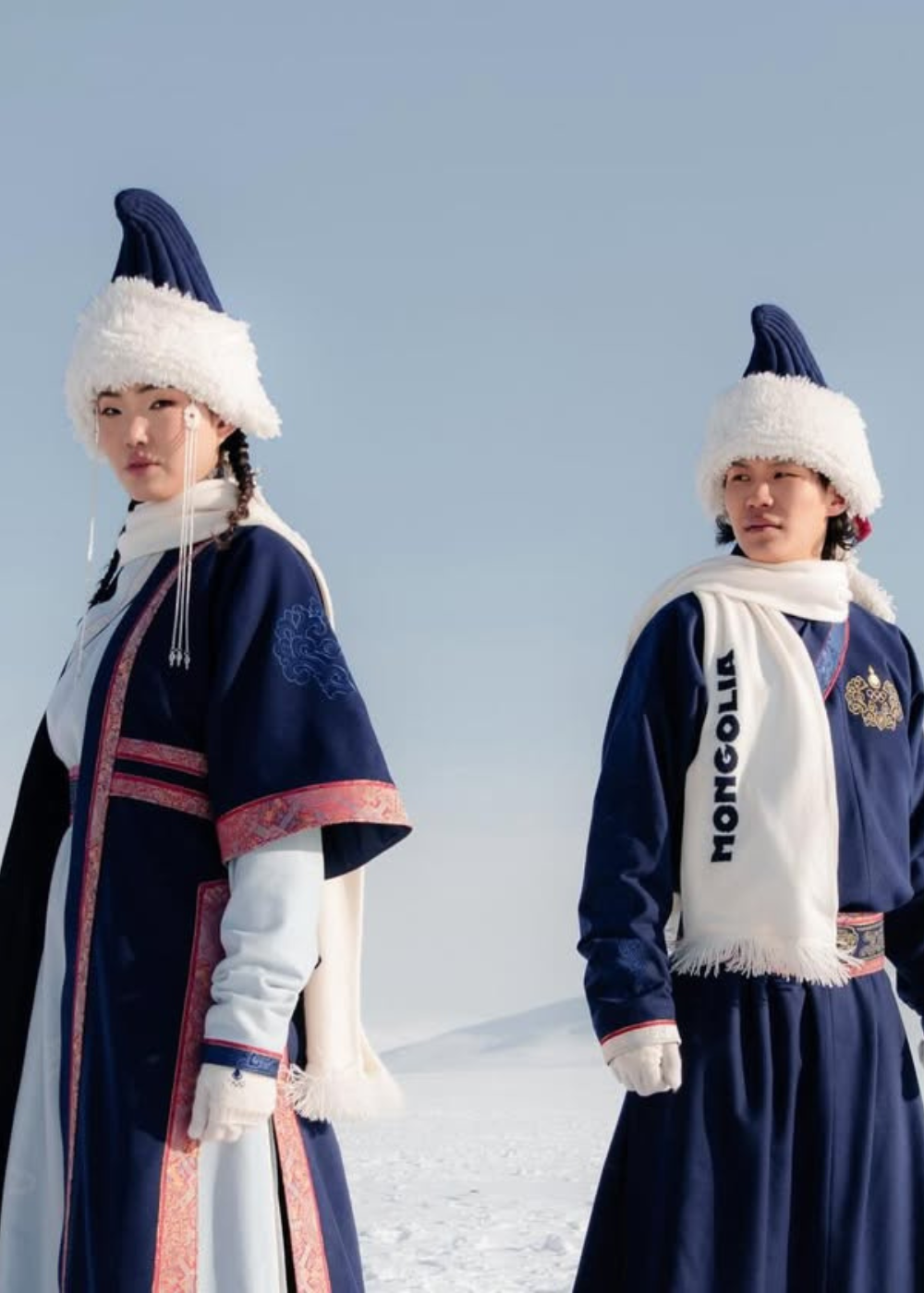 Dois modelos posam com trajes tradicionais mongóis em azul marinho e branco, com chapéus felpudos e cachecóis.