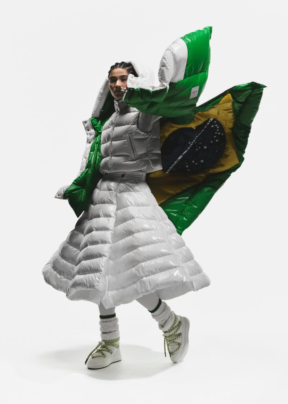Modelo veste casaco e saia plissada em nylon branco brilhante com botas e meias grossas, segurando capa verde com bandeira.