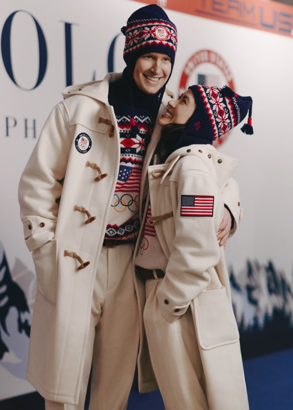 Casal abraçado veste casacos off-white com patch da bandeira dos EUA e gorros azul-marinho com padrão vermelho e branco.