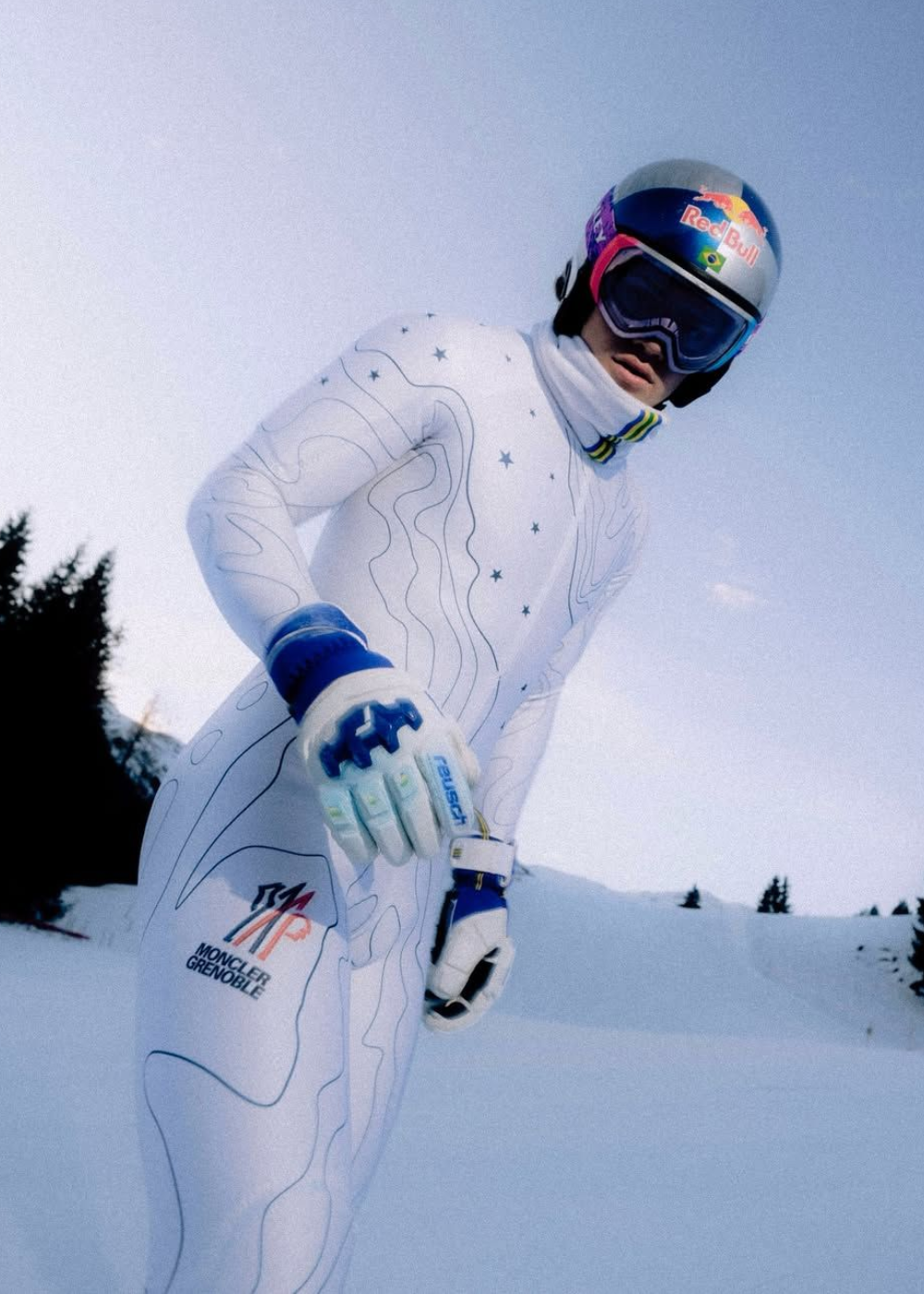 Atleta posa em macacão branco com estampas topográficas e estrelas, luvas e capacete prateado com detalhes azuis.