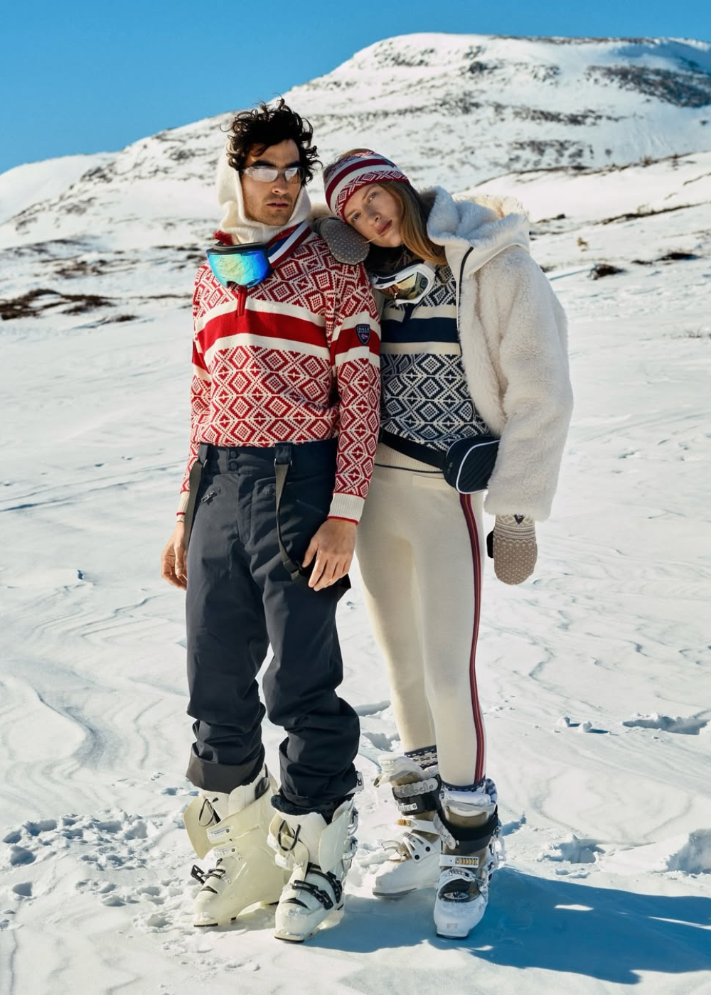 Casal posa junto em neve com suéteres de tricô geométrico vermelho e azul, calças térmicas e botas de esqui.