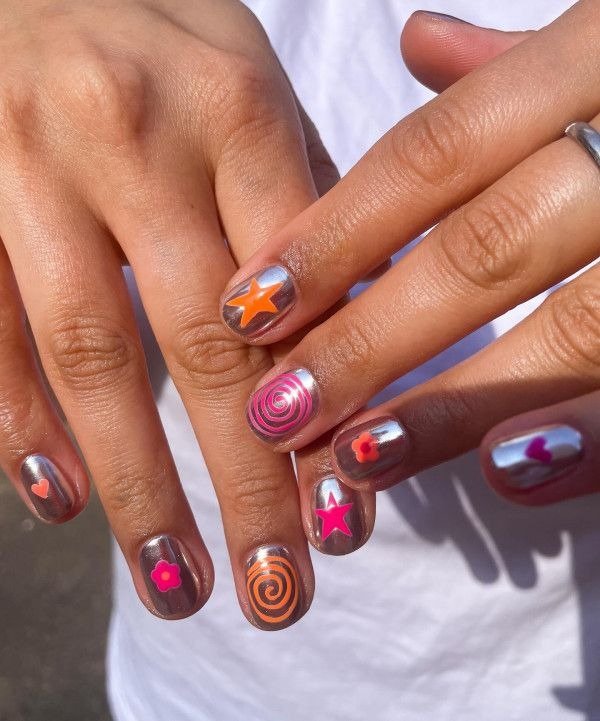 Unhas de verão com base metálica prateada e adesivos vibrantes em rosa, laranja e roxo, criando estilo divertido e moderno.