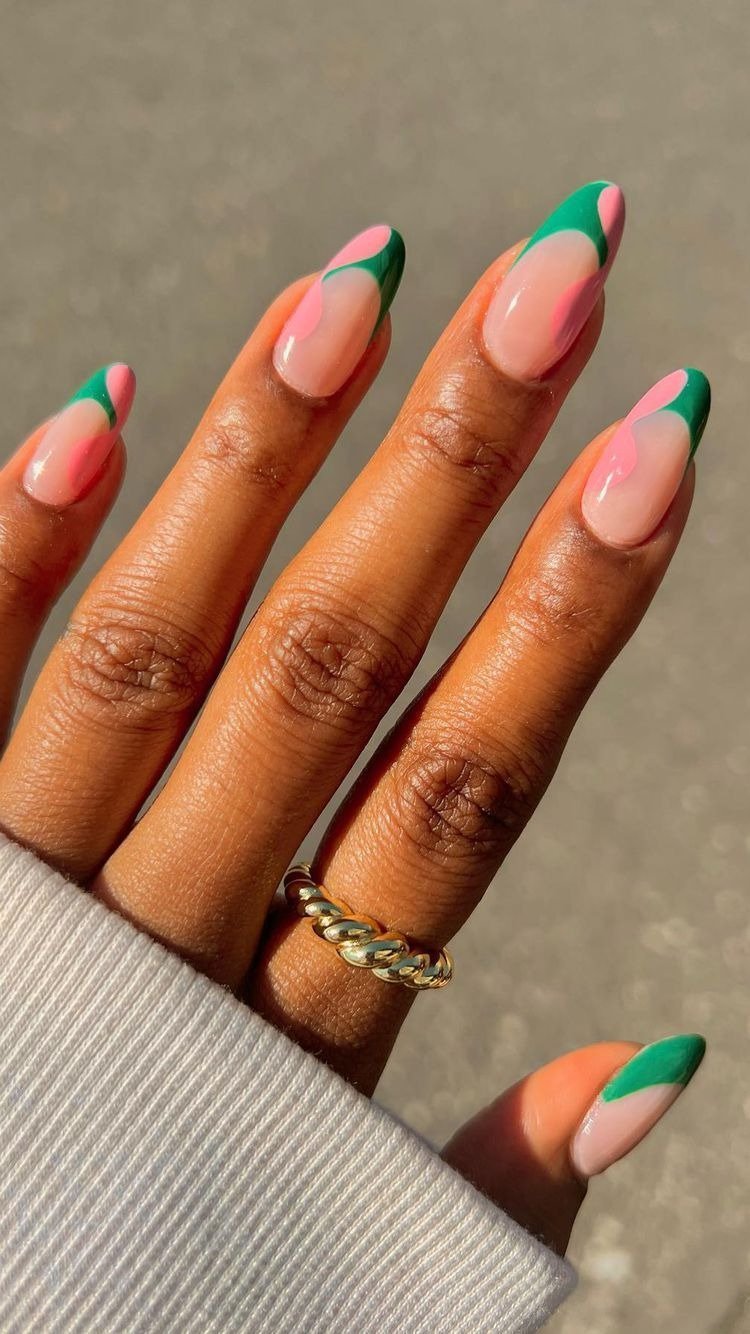 Unhas de verão com design abstrato em verde e rosa sobre base nude, formato amendoado e anel dourado delicado.