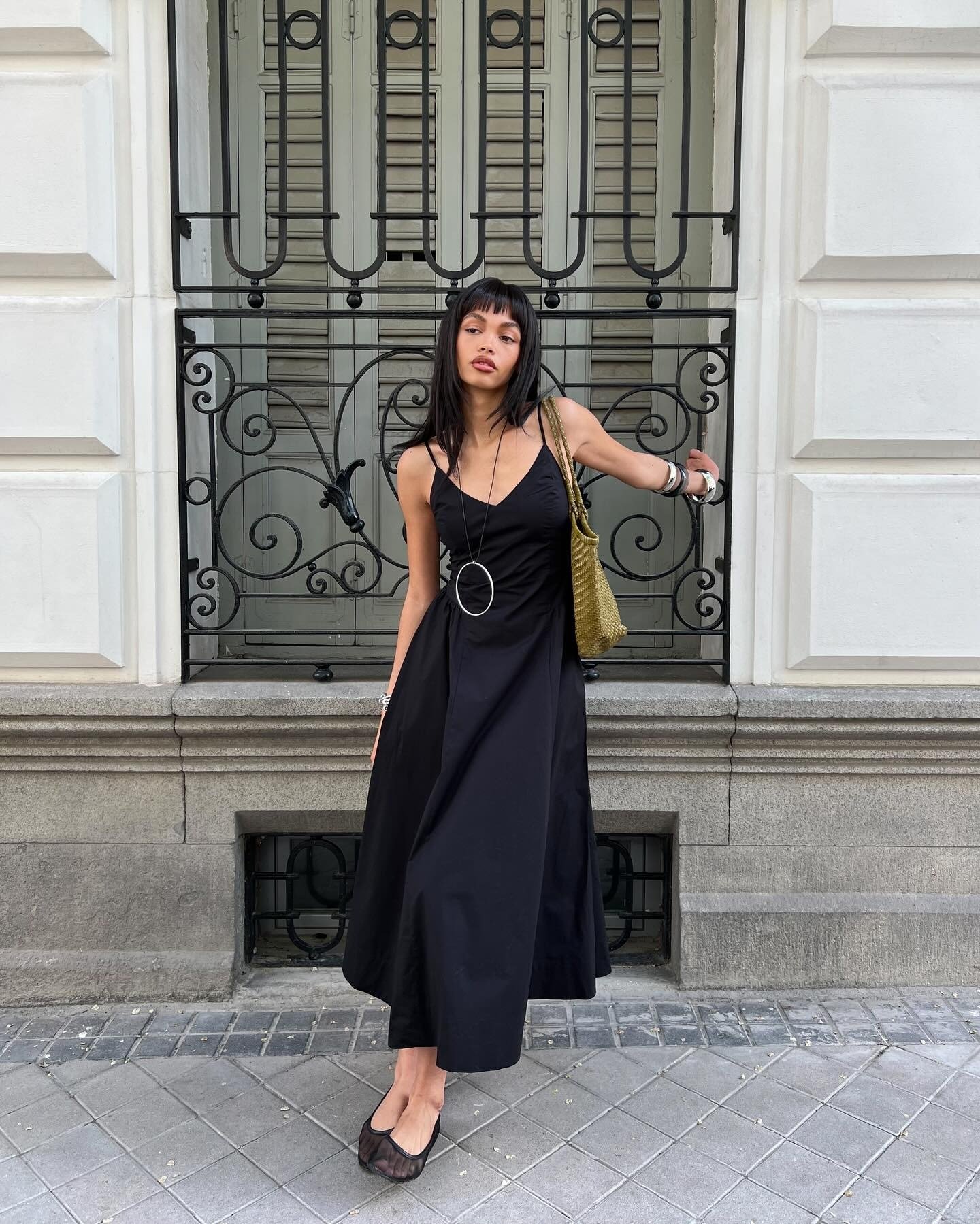 Modelo posa com vestido em crepe preto de alças finas e saia ampla, combinando acessórios prateados e bolsa verde musgo, em