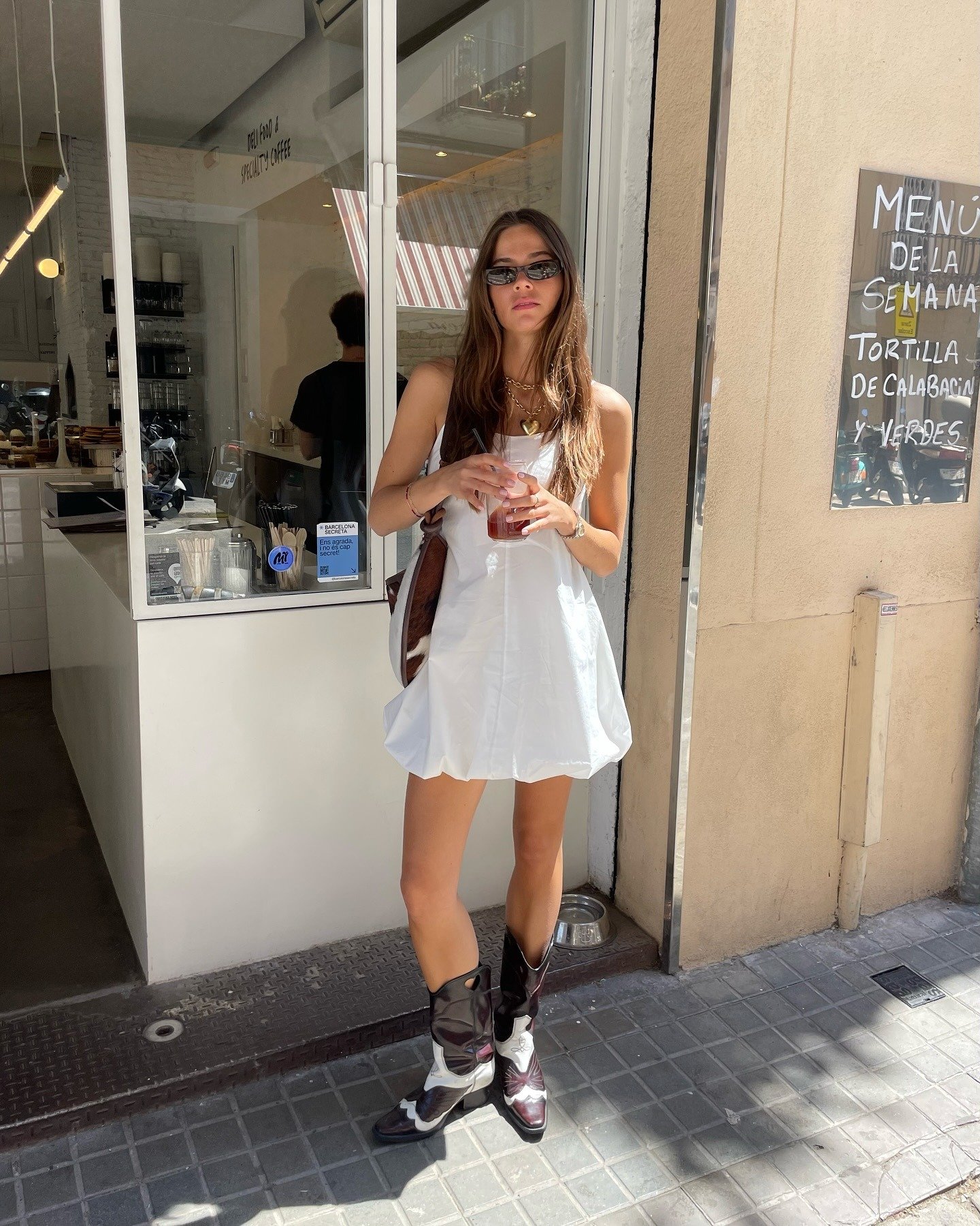 Modelo posa em vestido em crepe branco de modelagem soltinha com botas cowboy bicolor e acessórios dourados, em clima urbano