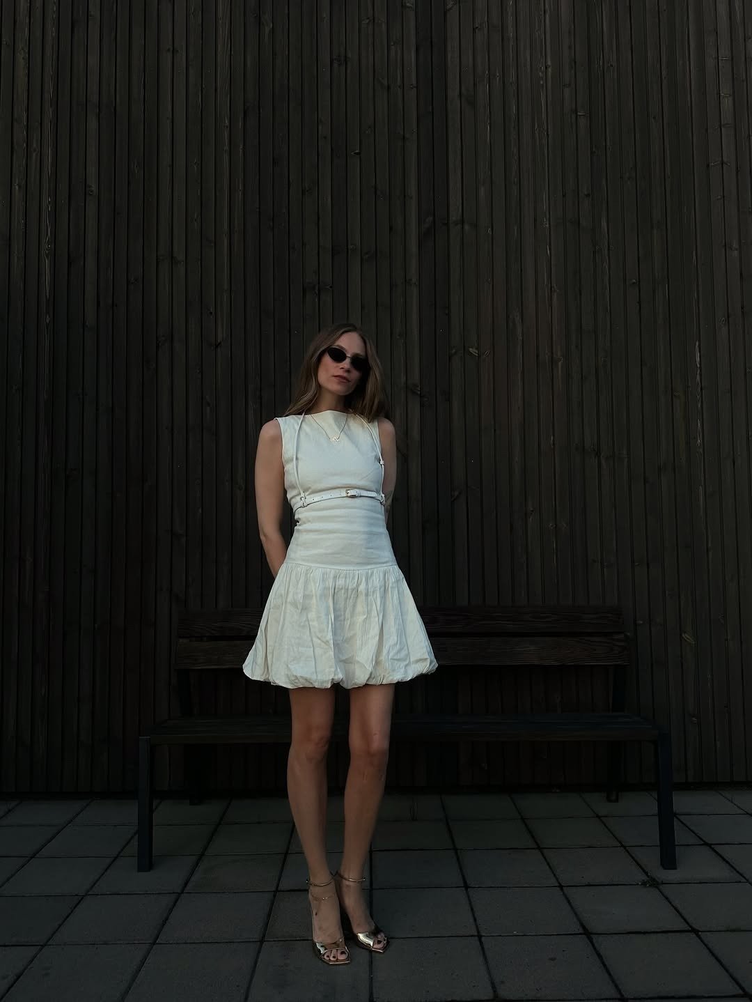 Modelo posa com vestido regata branco de tecido leve, saia bufante e cinto fino, combinando com sandálias douradas e óculos