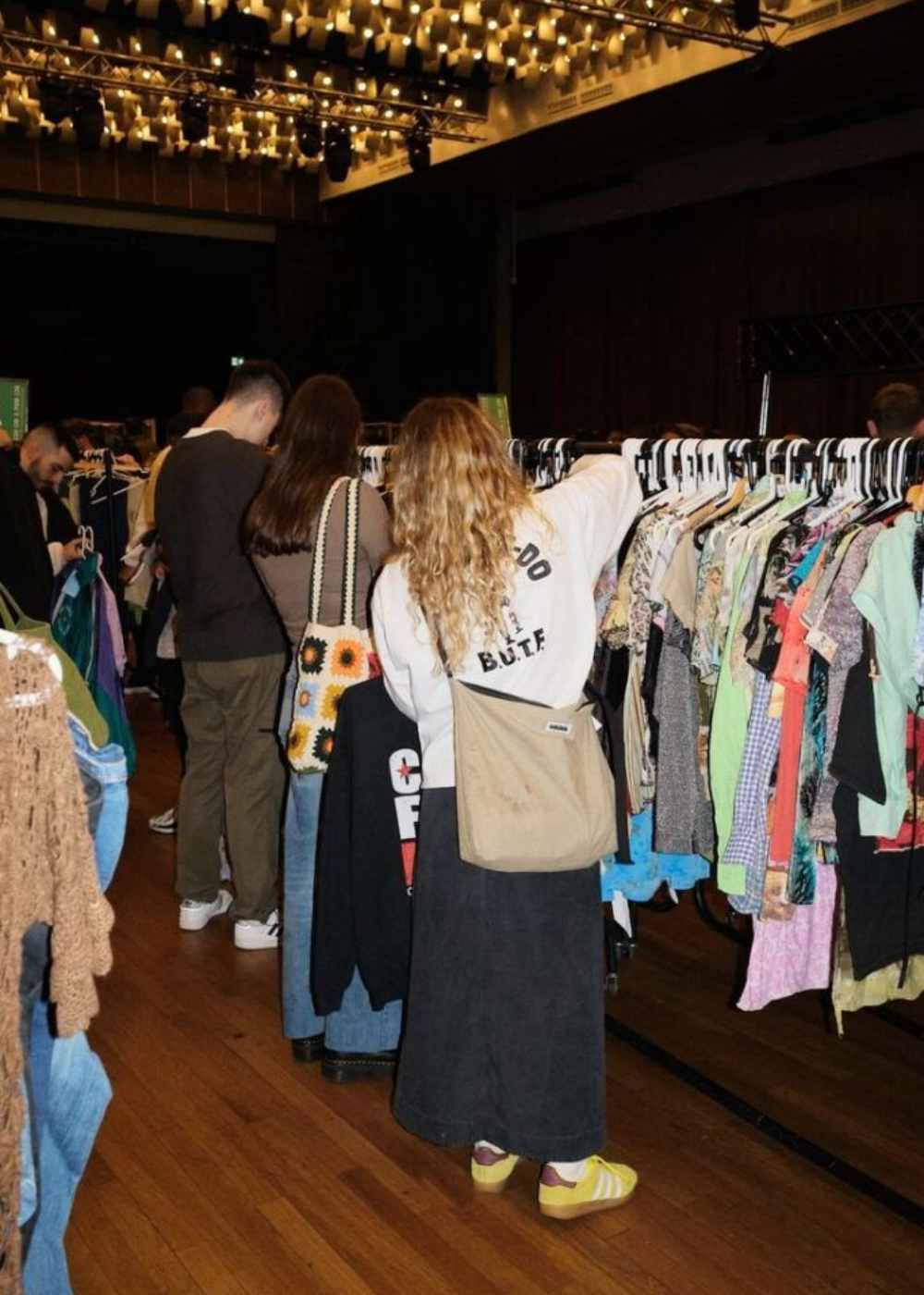 Pessoas exploram araras com tecidos variados, com modelagens soltas e cores neutras, em ambiente acolhedor e descontraído.