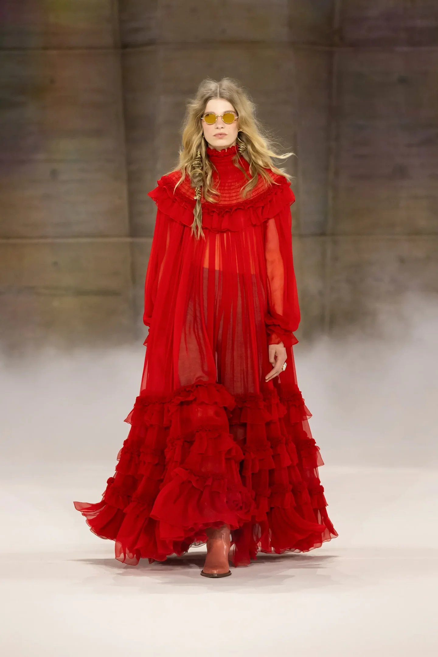 Modelo caminha com vestido longo vermelho de chiffon transparente, babados volumosos e gola alta, combinando botas e óculos