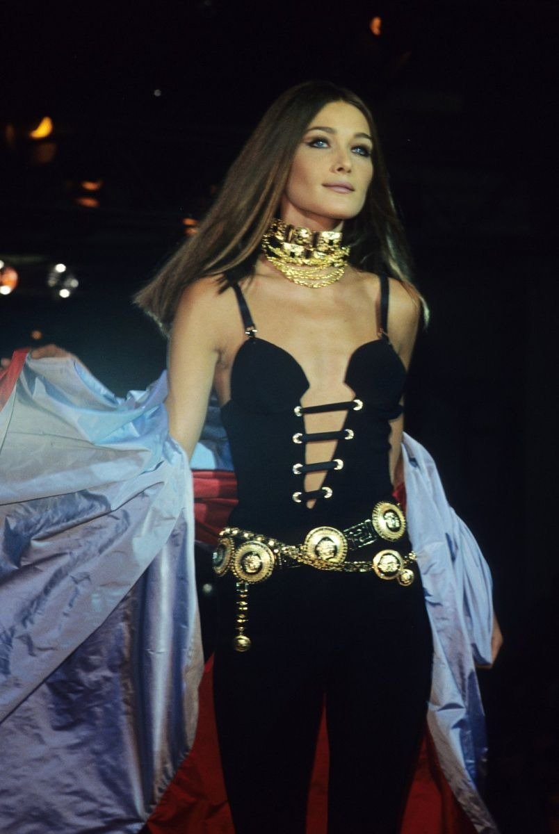 Modelo posa com macacão preto justo de decote ousado e tiras frontais, cinto e maxi colar dourados, em clima poderoso da