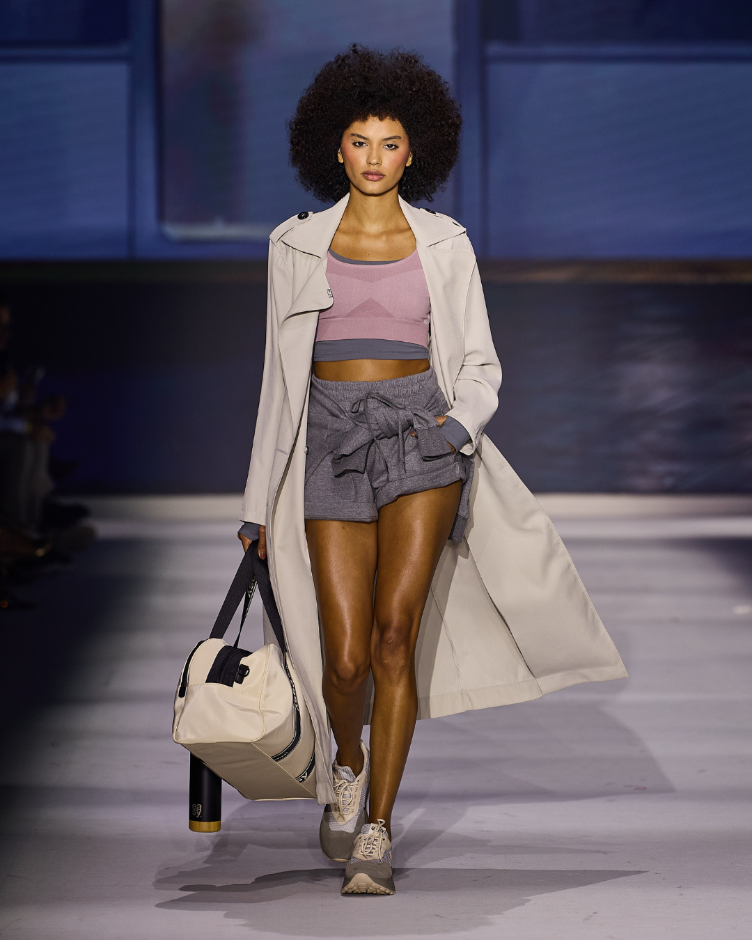 Tendências da primavera 2027: modelo desfila com trench bege, top rosa, short cinza e tênis, em clima esportivo elegante.