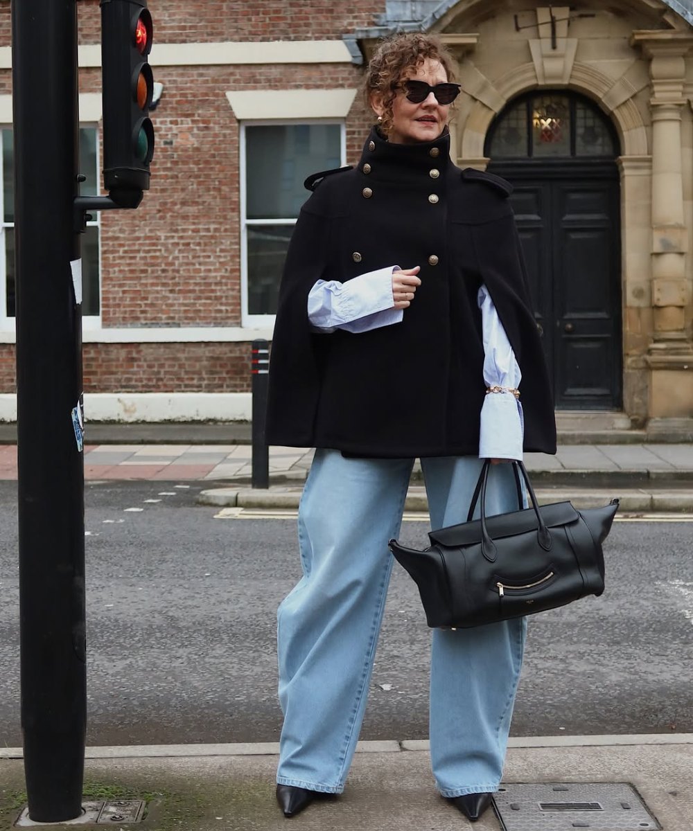 Modelo posa na rua com calça baggy jeans clara, capa preta estruturada, camisa branca e bolsa preta, em clima urbano.