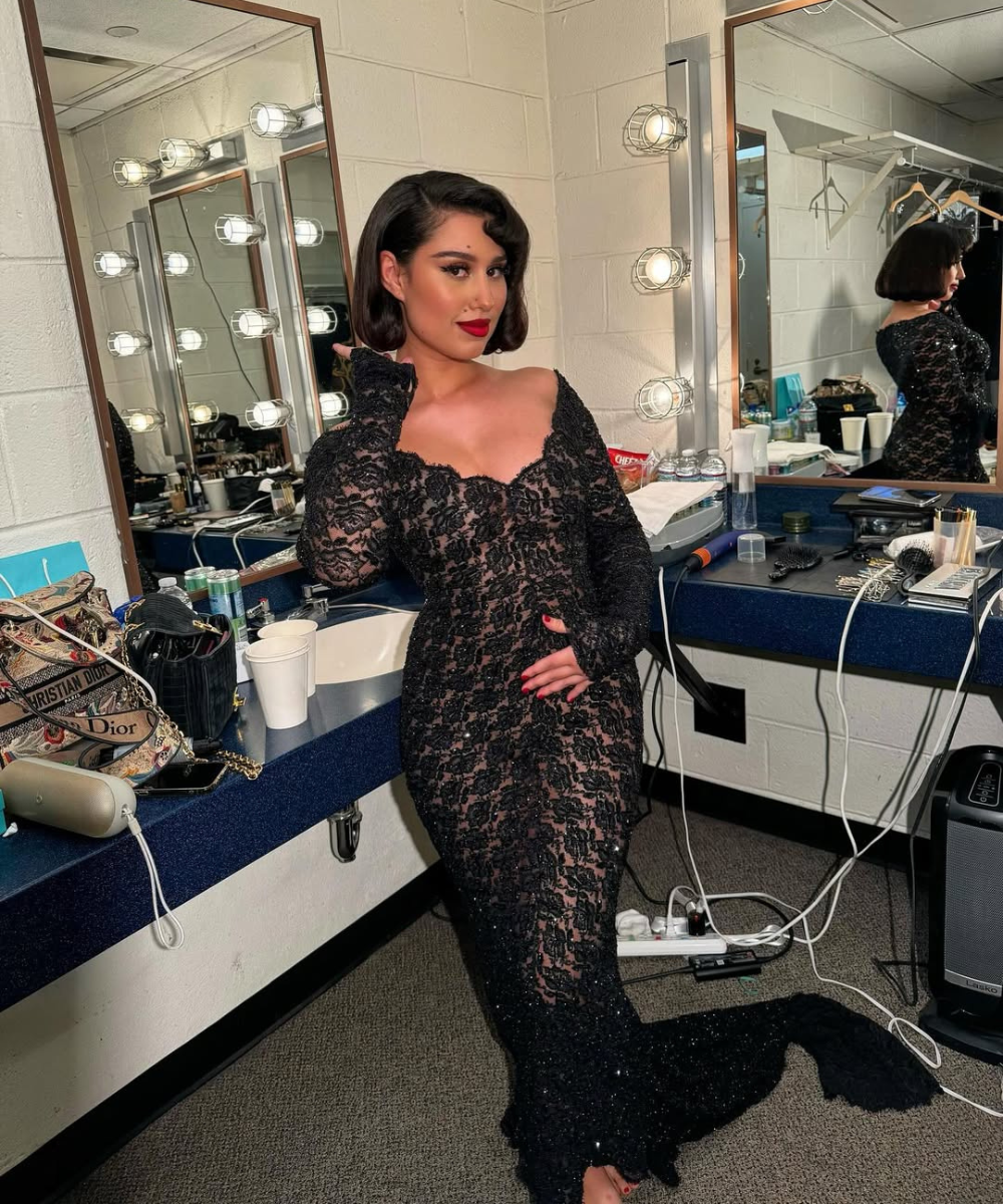 Escova modelada em cabelo curto, Raye posa com vestido sereia rendado preto em camarim iluminado.