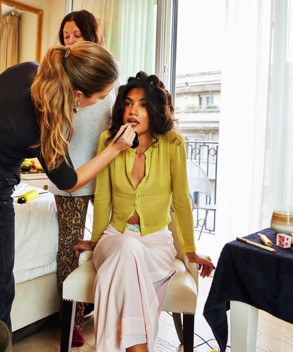 Escova modelada em cabelo cacheado; modelo posa sentada com blusa verde-limão e saia rosa enquanto recebe maquiagem.