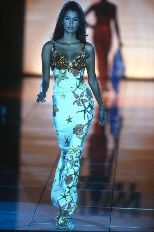 Modelo caminha com postura confiante usando vestido longo Versace justo em cetim branco, estampas marinhas e detalhes