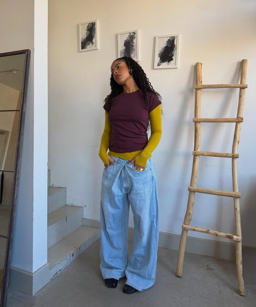 Modelo posa com calça baggy jeans clara, blusa vinho e manga mostarda.