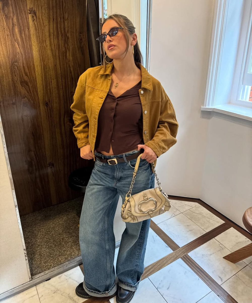 Modelo posa com calça baggy jeans azul, jaqueta caramelo blusa e cinto marrom em look urbano sofisticado e confiante.