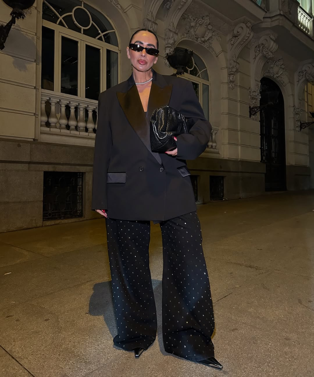 Modelo posa à noite com calça baggy preta de brilho sutil, blazer amplo e clutch, em clima urbano sofisticado.