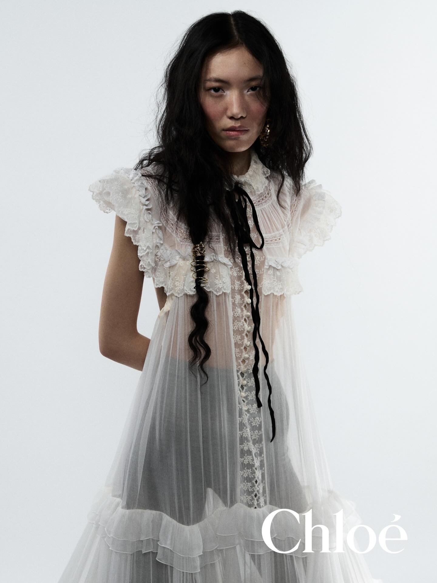 Modelo posa com vestido branco translúcido de tule e renda, babados delicados e laço preto no pescoço, expressando elegância