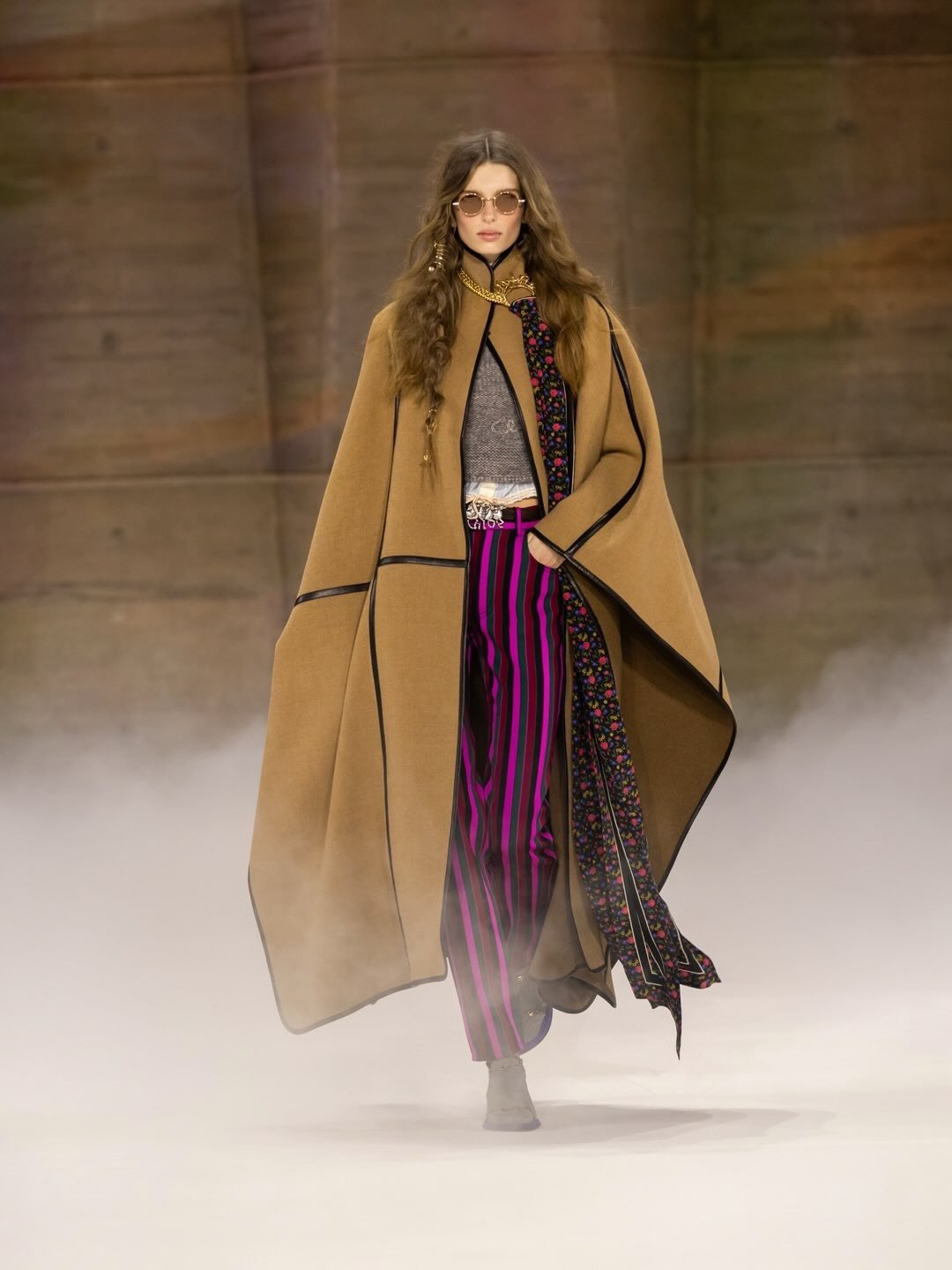 Modelo da Chloé caminha confiante usando capa camel com acabamento preto, blusa cropped cinza e calça listrada rosa e preta,