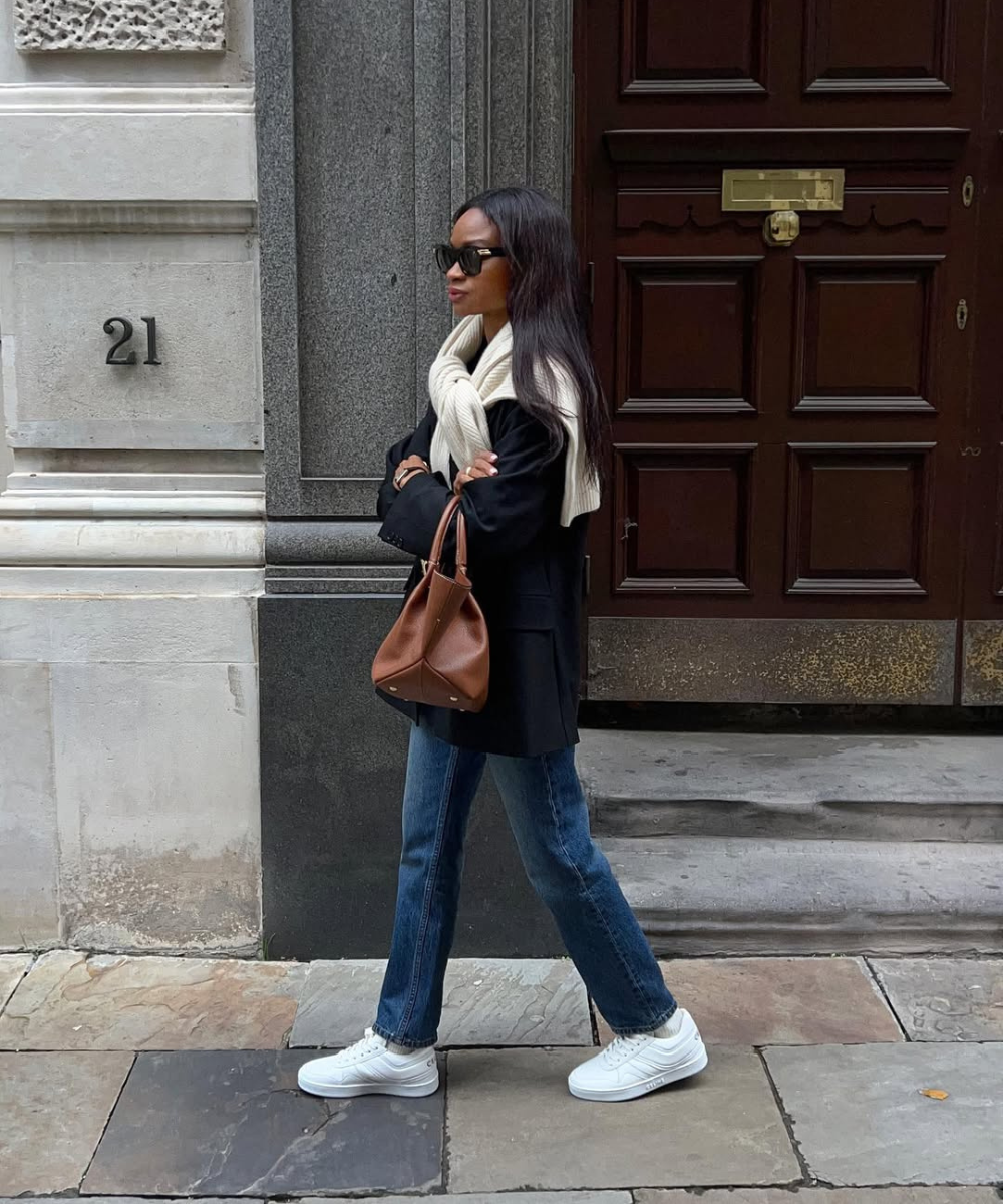 Looks com jeans para usar no trabalho: modelo caminha com blazer preto, jeans reto azul, tricô creme e bolsa marrom.