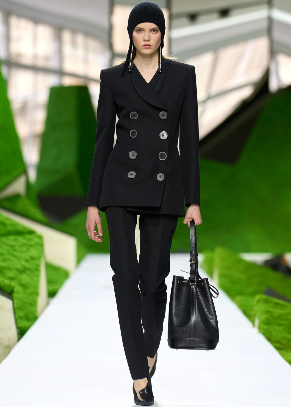 Outono/Inverno 2026: modelo posa em alfaiataria preta acinturada, gorro canelado e bolsa estruturada, com sobriedade urbana.