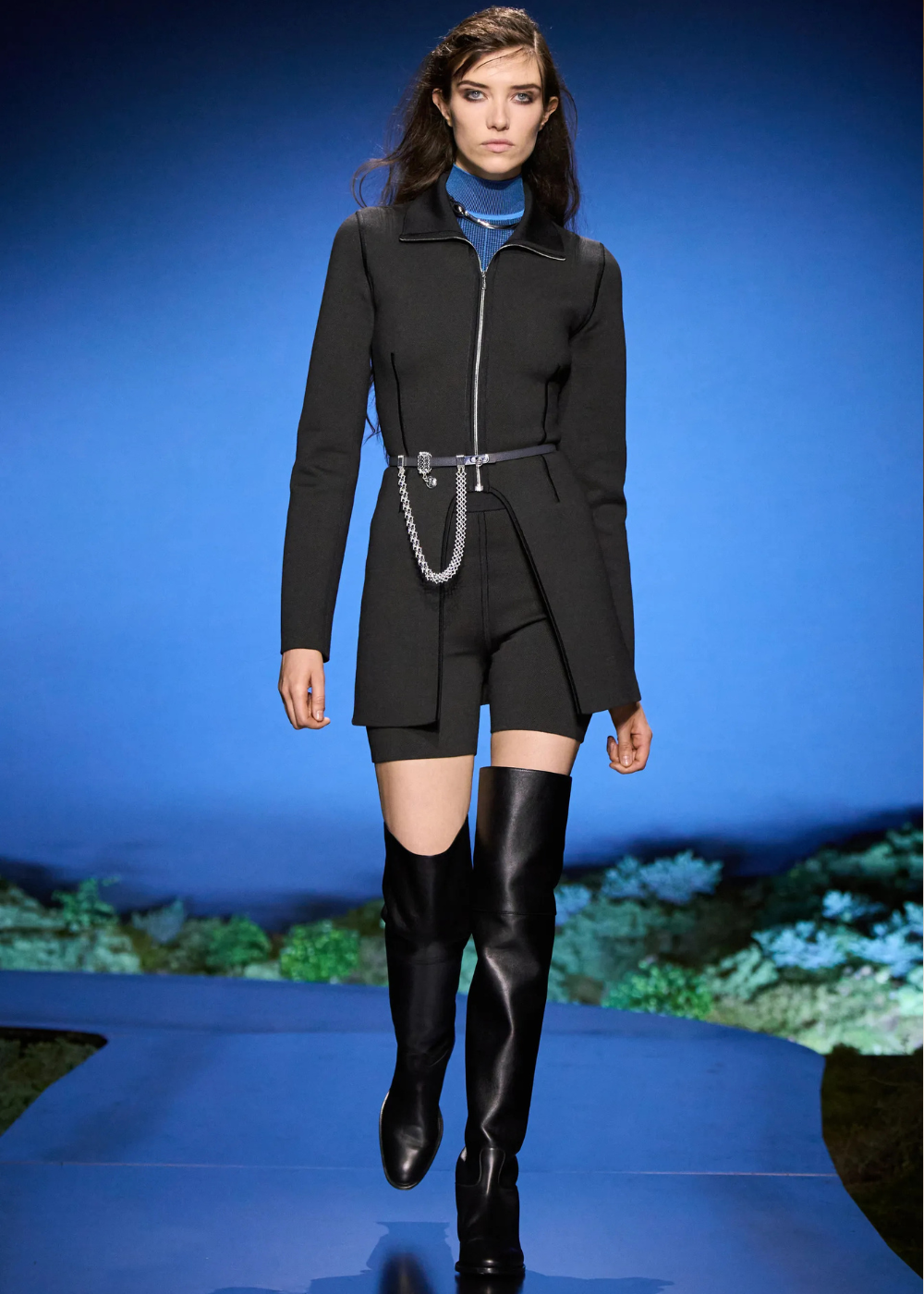 Outono/Inverno 2026: modelo desfila look preto ajustado com zíper, gola azul e bota over the knee em clima futurista.