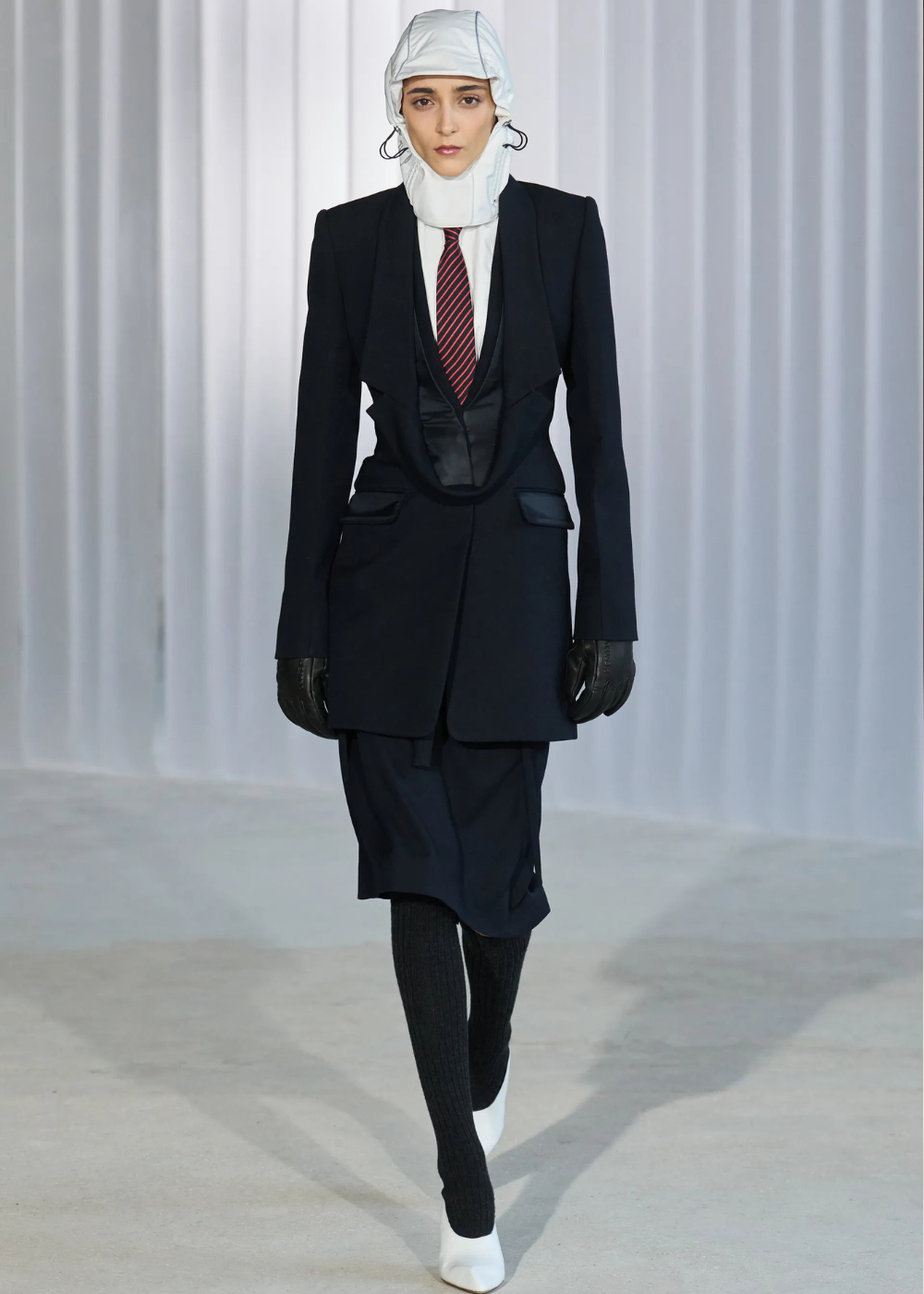 Outono/Inverno 2026: modelo desfila de blazer preto estruturado, gravata listrada e capuz branco em clima austero.