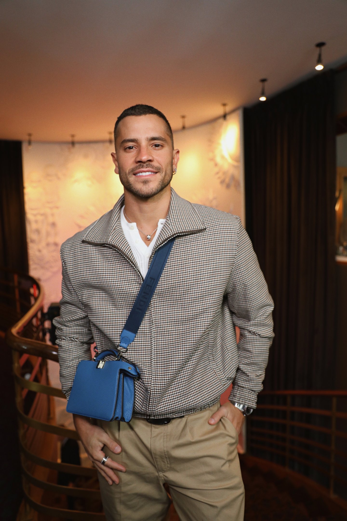 Celebs exibem estilo casual com jaqueta xadrez, calça bege e bolsa azul Fendi, posando confiante em ambiente sofisticado.