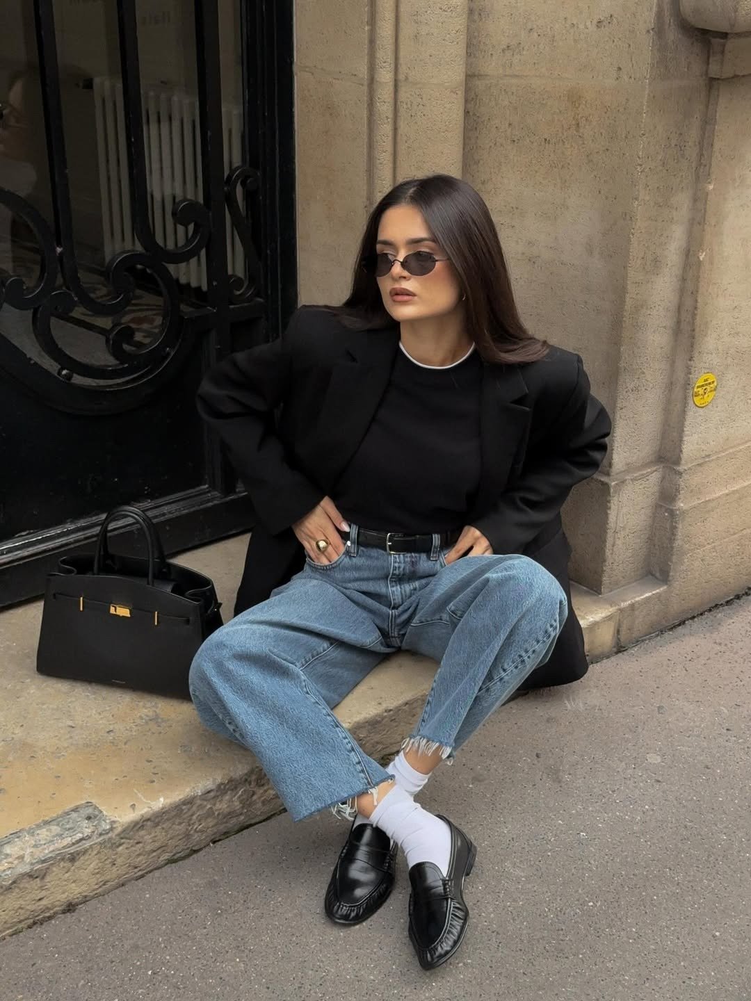 Mocassim feminino com calça jeans reta, blazer preto e meia branca; modelo posa sentada na calçada com elegância urbana.