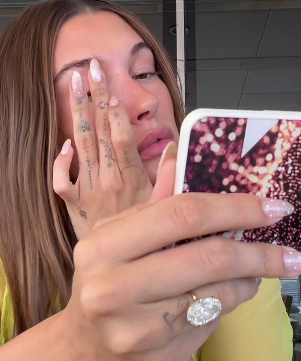 Modelo posa de amarelo com unhas claras e anel de noivado oval brilhante enquanto retoca a maquiagem ao espelho do celular