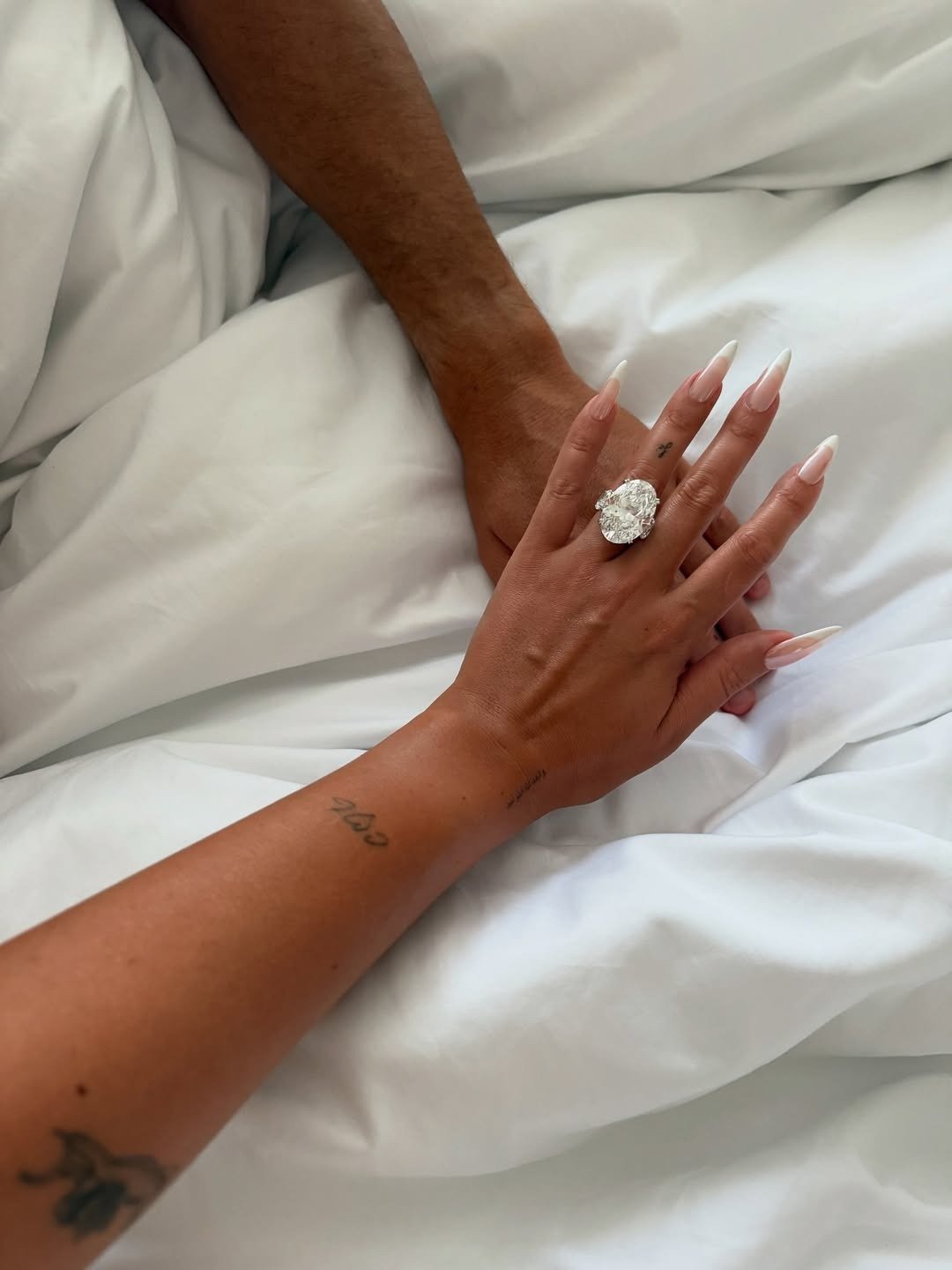 Anel de noivado oval com diamante maxi brilha em mão com unhas nude sobre lençóis brancos, em clima íntimo.
