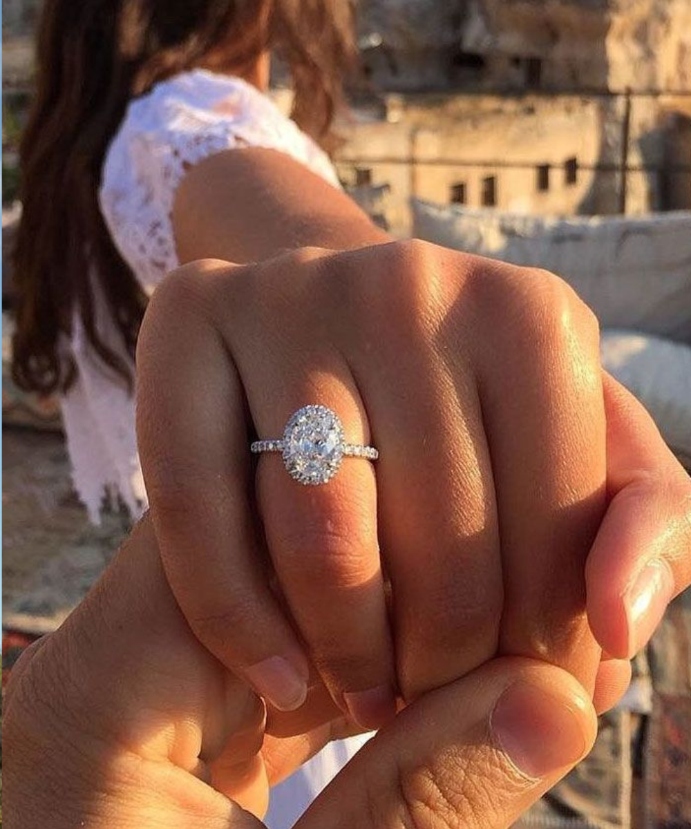 Anel de noivado oval em ouro branco com halo brilhante destaca mão estendida, vestido rendado branco e clima romântico