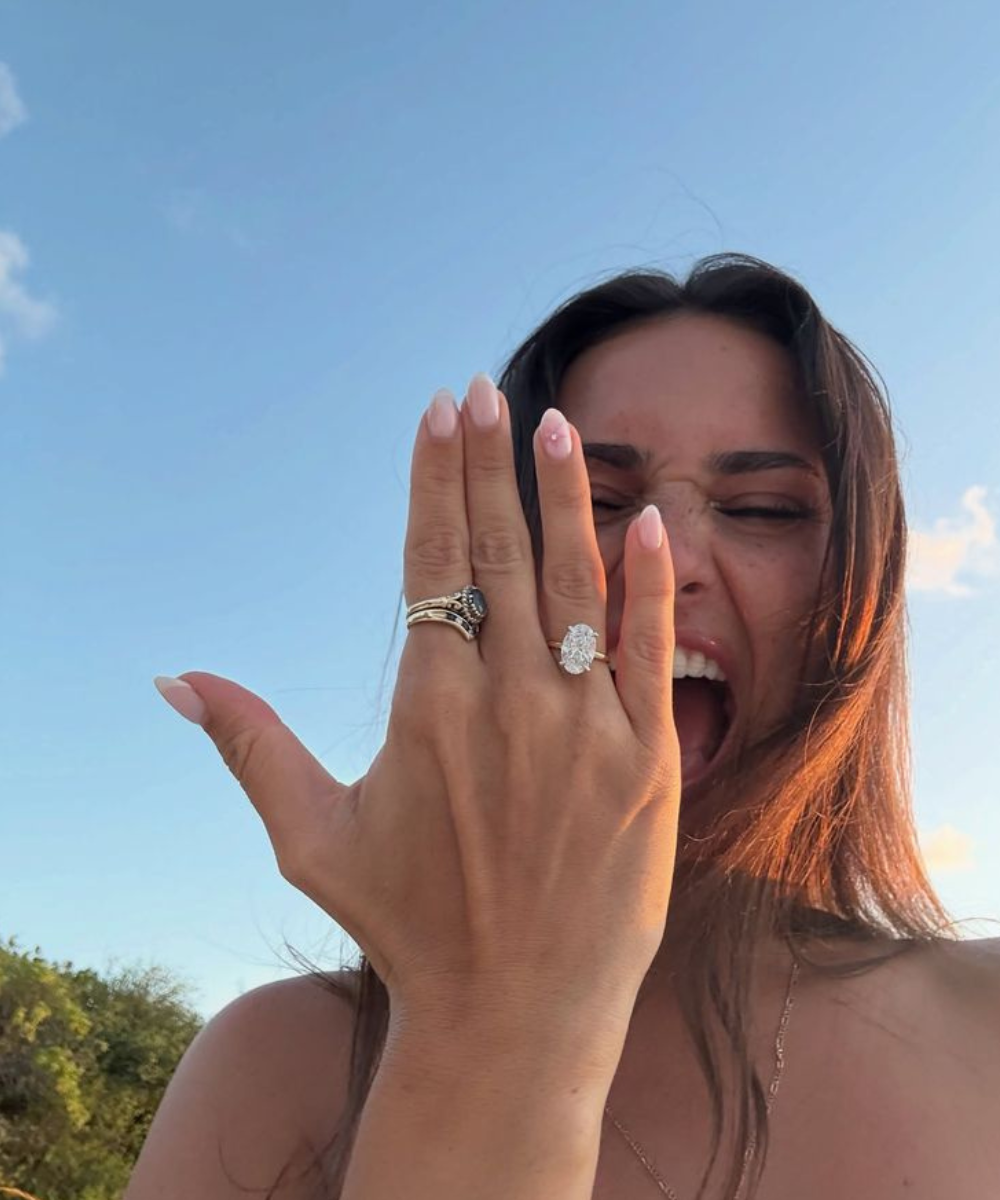 Modelo posa ao ar livre com anel de noivado oval dourado, unhas nude e expressão radiante sob céu azul.