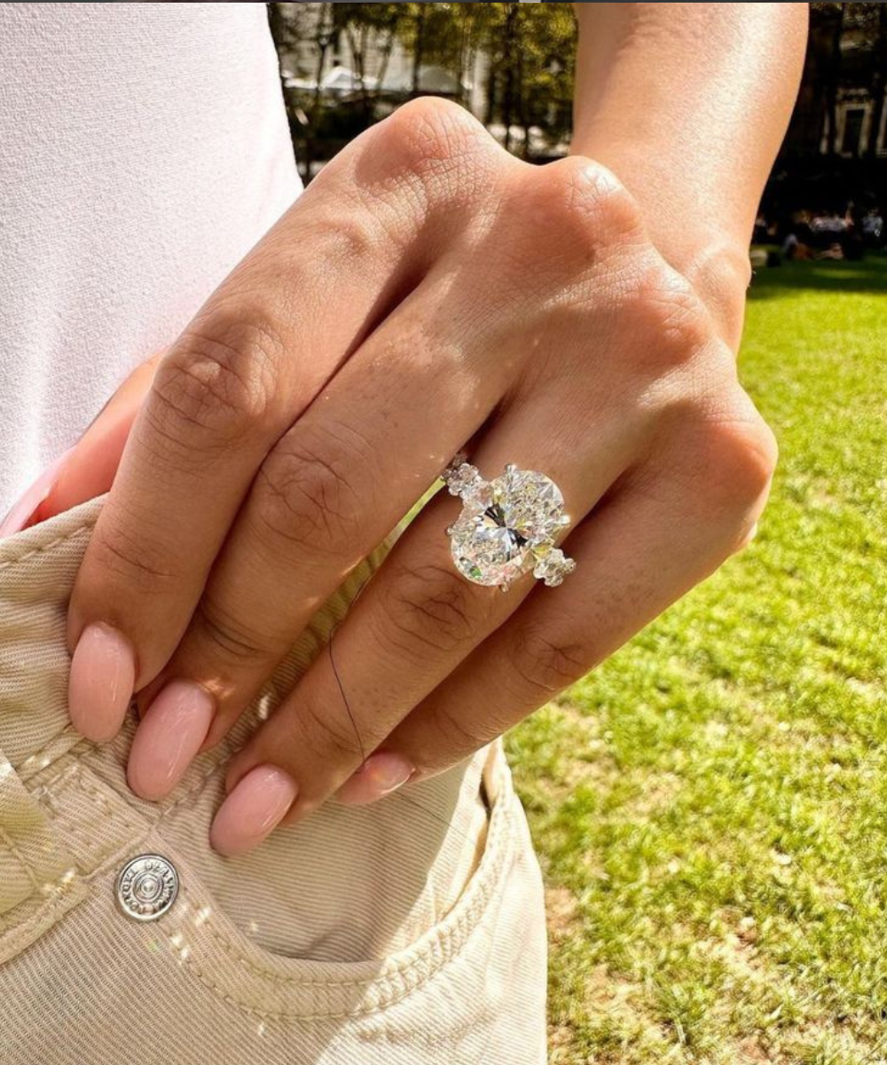 Anel de noivado oval com diamante brilhante em mão com unhas nude, calça bege e gramado ao sol, clima elegante e romântico