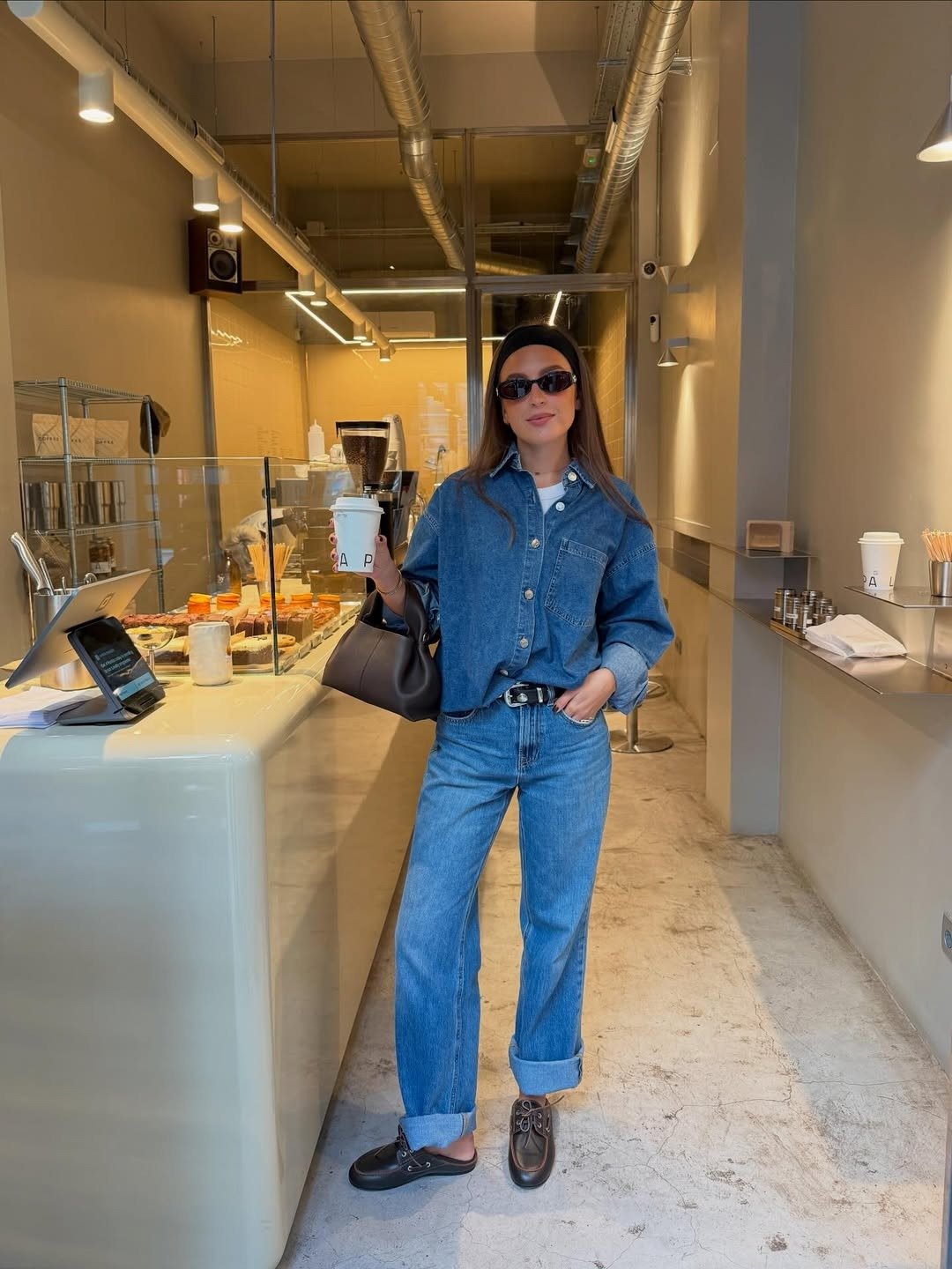 Modelo posa com mocassim feminino com calça jeans reta e camisa denim azul em café moderno, com elegância casual.