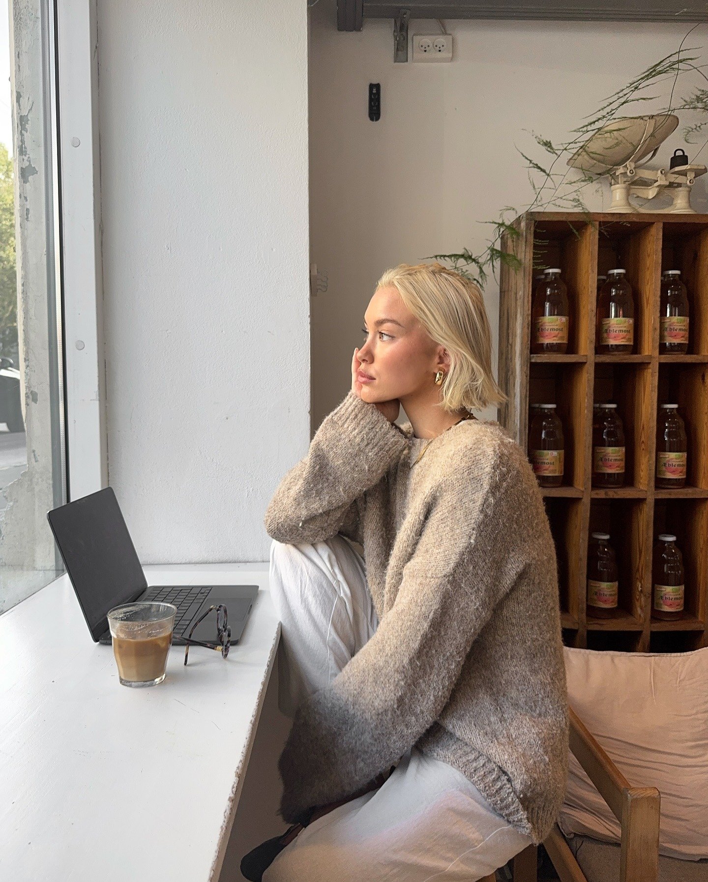 Ansiedade em café: modelo posa de perfil com tricô oversized bege e calça branca, em clima introspectivo junto ao laptop.