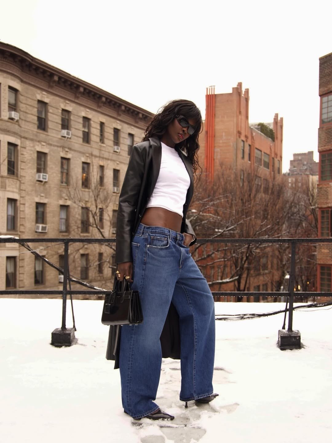 Mulher na neve com look de peças de ármario capsula. O look é composto por blazer preto, cropped branco, jeans amplo e bolsa